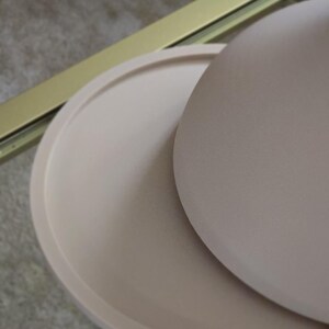 May include: A set of two round, beige ceramic trays. The larger tray is slightly domed and sits on top of the smaller, flat tray.