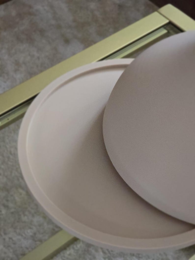May include: A set of two round, beige ceramic trays. The larger tray is slightly domed and sits on top of the smaller, flat tray.