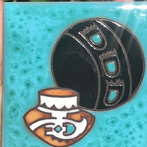 May include: A turquoise ceramic tile with a black and white design of a pot and a ball. The pot is brown with white accents and a turquoise design. The ball is black with white accents and turquoise designs.