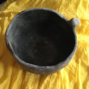 以下が含まれることがあります： 黄色の布の上に置かれた、単一の取っ手を持つ濃い灰色のセラミックボウル