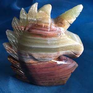 Puede incluir: Una escultura de unicornio de ónix tallado. La escultura presenta una melena y un cuerno detallados, con capas de colores verde, marrón y crema. La base es de un marrón más oscuro. La escultura está sobre un fondo azul.