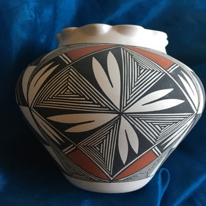 Puede incluir: Una olla de cerámica blanca con un diseño geométrico en negro y naranja. La olla tiene un borde festoneado y está decorada con un patrón repetitivo de triángulos y hojas.