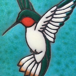 May include: A ceramic tile with a painted image of a hummingbird with green wings, a white body, and a red throat. The hummingbird is flying with its wings spread out. The background is a turquoise blue.