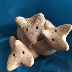 以下が含まれることがあります： 3匹の小さな茶色の粘土の狐が寄り添っています。狐は小さな黒い目をしていて、ざらざらとした質感の粘土でできています。