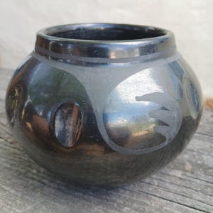 Könnte beinhalten: Ein schwarzer Keramiktopf mit rundem Körper und breiter Öffnung. Der Topf hat eine glänzende Oberfläche und ein mattes Vogeldesign. Der Topf steht auf einer Holzoberfläche.