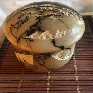 Puede incluir: Una pequeña caja redonda de madera con tapa. La caja está decorada con un diseño en blanco y negro que se asemeja a una cordillera. La caja está sentada sobre una estera tejida marrón.