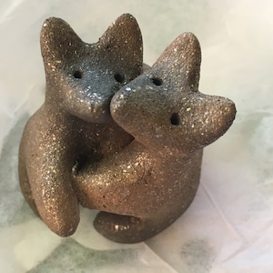 以下が含まれることがあります： 斑点模様の茶色の陶器製のキツネが2匹、向き合っています。キツネは遊び心のあるポーズで彫刻されており、1匹のキツネの頭がもう1匹のキツネの背中に乗っています。