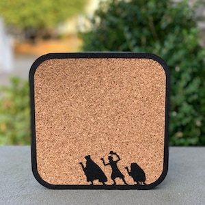 May include: A corkboard with a black frame and a silhouette of three figures from the Haunted Mansion attraction. The figures are a man in a top hat, a woman with a hat, and a dog.
