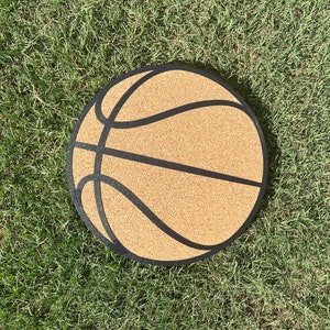 Puede incluir: Un tablero de corcho con forma de balón de baloncesto con un contorno negro. El tablero de corcho está sobre una superficie de hierba verde.