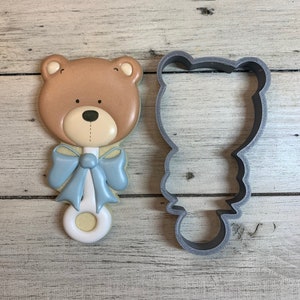 May include: A brown bear-shaped cookie cutter with a blue bow and a white handle. The cookie cutter is next to a decorated cookie in the same shape.