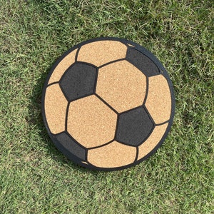 Puede incluir: Una alfombra de bienvenida redonda de corcho y negro con diseño de balón de fútbol. La alfombra tiene un fondo de corcho marrón con costuras negras de balón de fútbol.