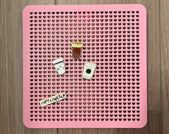 May include: A pink pegboard with a heart-shaped grid pattern. There are four enamel pins on the board, each depicting a different type of coffee drink. One pin says "caffeinated" in black and white text.