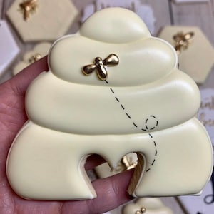 May include: A white sugar cookie decorated to look like a beehive with a gold bee flying towards it. The cookie has a black outline and a small opening at the bottom.