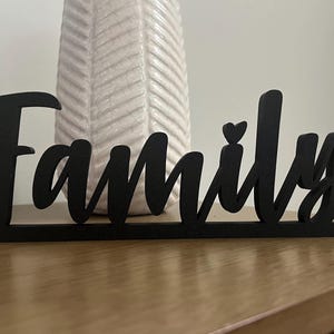 May include: Black wooden sign with the word "Family" in a decorative font. A small heart is included in the word.