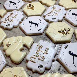 May include: A collection of decorated sugar cookies with a bee theme. The cookies are decorated with gold and black icing and feature the phrase "Meant to bee" and a honeycomb pattern.