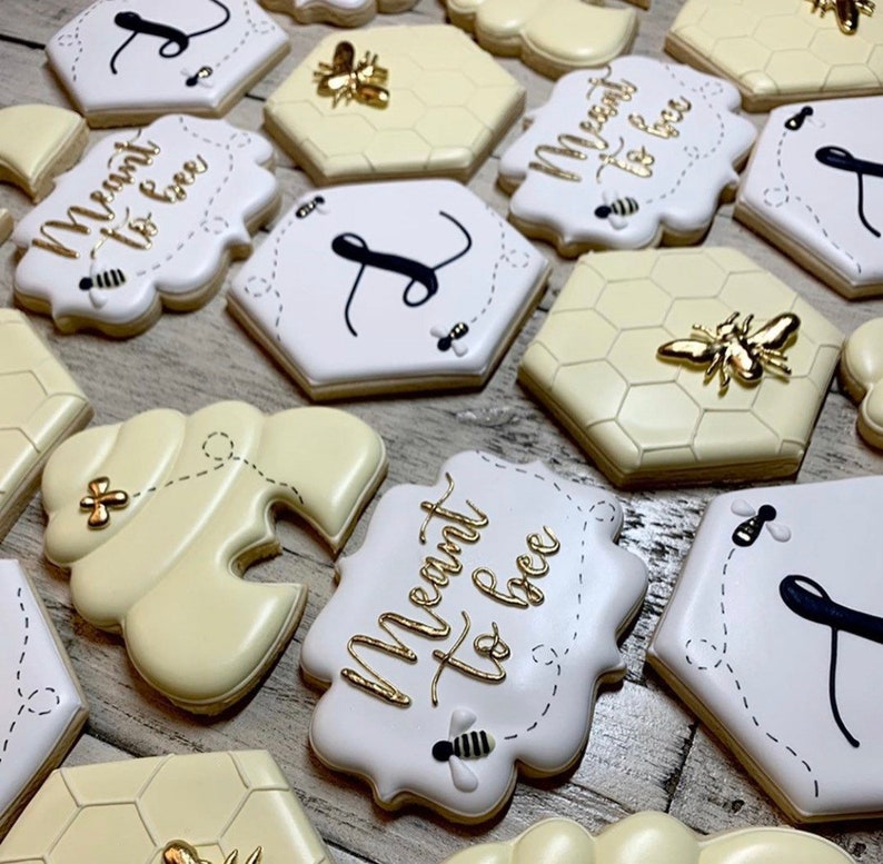 May include: A collection of decorated sugar cookies with a bee theme. The cookies are decorated with gold and black icing and feature the phrase "Meant to bee" and a honeycomb pattern.