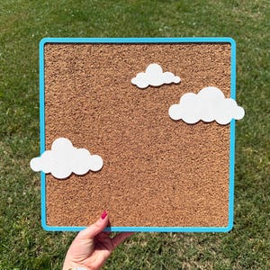 May include: A square corkboard with a light blue frame and three white cloud cutouts. The corkboard is held up against a background of green grass.