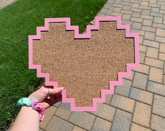May include: A pink heart-shaped cork board with a pixelated border. The cork board is made of natural cork and is perfect for pinning notes, photos, or other items.