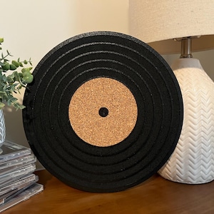 May include: Black vinyl record coaster with a cork centre. The coaster is round and has a textured surface.