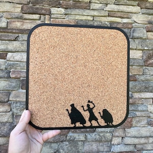 May include: A cork board with a black frame and a silhouette of four figures, including a ghost, a witch, and two other figures. The board is made of cork and has a brown color.