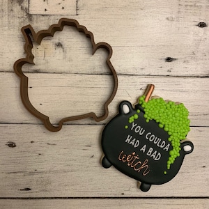 Könnte beinhalten: Ein schwarzer und grüner Keks in Form eines Kessels mit dem Text "You coulda had a bad witch" darauf. Der Keks liegt neben einem braunen Ausstecher in Form eines Kessels.