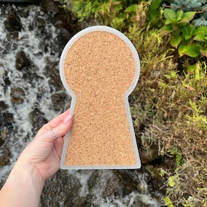 May include: A cork coaster shaped like a keyhole. The coaster is gray with a cork center.