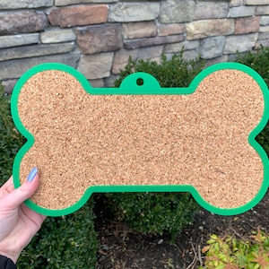 Puede incluir: Un tablero de corcho con forma de hueso de color verde con una superficie de corcho. El tablero tiene un agujero en la parte superior para colgarlo.