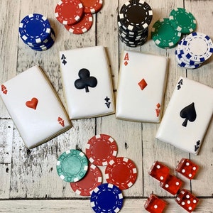 May include: A set of four decorated sugar cookies shaped like playing cards. The cookies are decorated with red, black, and white icing. The cookies are arranged on a white wooden surface with various poker chips and playing cards.