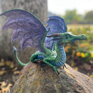 Op de afbeelding: Gedetailleerd beeldje van een draak met groene schubben, een blauwe buik en paarse vleugels. De draak zit op een rots, met zijn vleugels gespreid. De draak heeft een lange staart en kijkt opzij.
