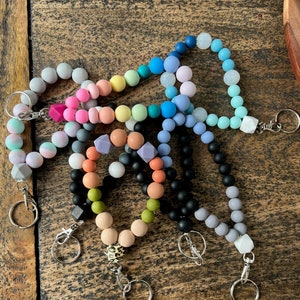 May include: A collection of beaded wristlet keychains in various colors, including gray, pink, blue, and black. Each keychain features a silver clasp and key ring. Some keychains have geometric or patterned beads.
