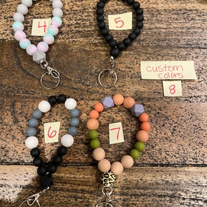May include: A collection of beaded wrist keychains in various colors. The keychains feature round beads in colors like black, gray, pink, and orange. Some keychains have a leopard print accent bead. The image includes a note that says "custom colors".