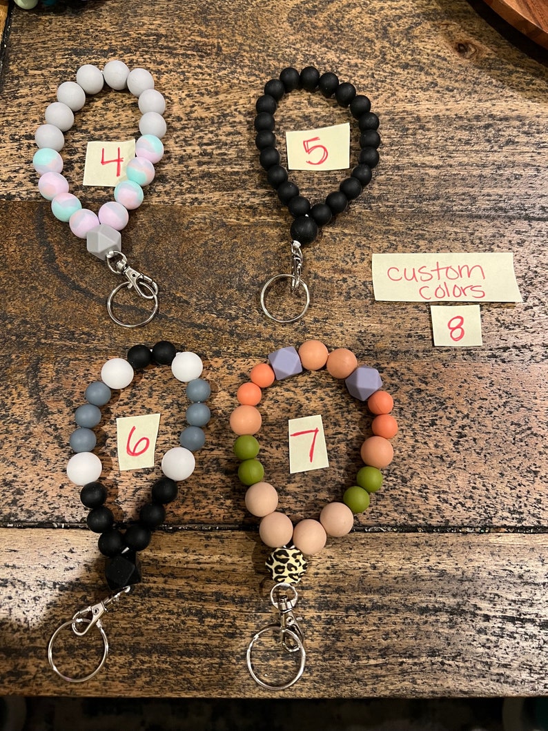 May include: A collection of beaded wrist keychains in various colors. The keychains feature round beads in colors like black, gray, pink, and orange. Some keychains have a leopard print accent bead. The image includes a note that says "custom colors".