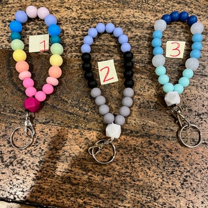 May include: Three beaded wristlet keychains in a variety of colors. Keychain 1 features a rainbow of colors, keychain 2 has black, gray, and lavender beads, and keychain 3 has blue and white beads. Each keychain has a silver clasp and a small tag with a number.