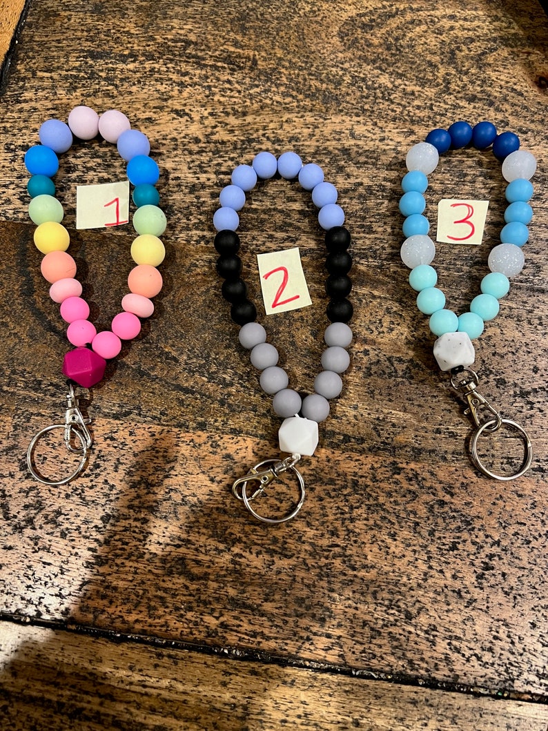 May include: Three beaded wristlet keychains in a variety of colors. Keychain 1 features a rainbow of colors, keychain 2 has black, gray, and lavender beads, and keychain 3 has blue and white beads. Each keychain has a silver clasp and a small tag with a number.