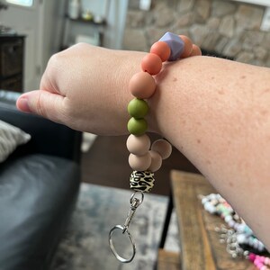 May include: A wristlet keychain with a variety of colorful silicone beads in shades of peach, green, and tan. The keychain includes a leopard print bead and a silver-colored clasp and key ring.