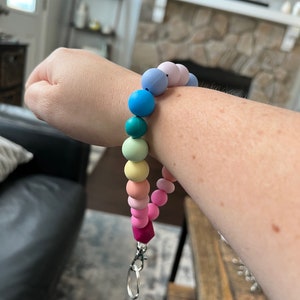May include: A rainbow-colored silicone bead wristlet with a silver-colored clasp. The wristlet features a variety of pastel-colored beads, including pink, yellow, green, blue, and purple. The wristlet is designed to hold keys or other small items.
