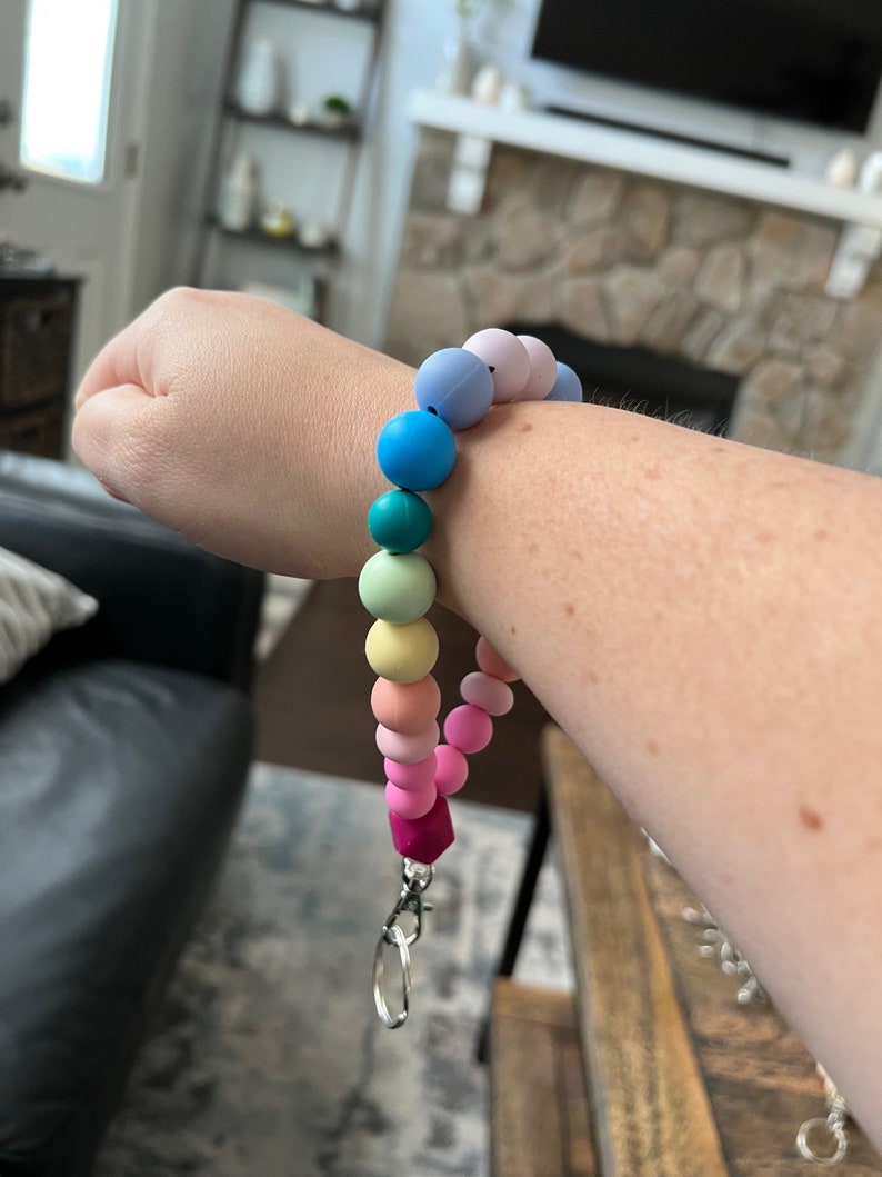 May include: A rainbow-colored silicone bead wristlet with a silver-colored clasp. The wristlet features a variety of pastel-colored beads, including pink, yellow, green, blue, and purple. The wristlet is designed to hold keys or other small items.