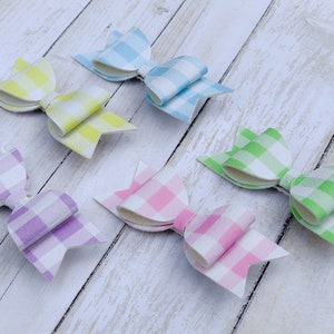 May include: Five gingham hair bows in different pastel colours. The bows are made of fabric with a white and coloured checkered pattern. The colours are light blue, yellow, green, pink, and purple.