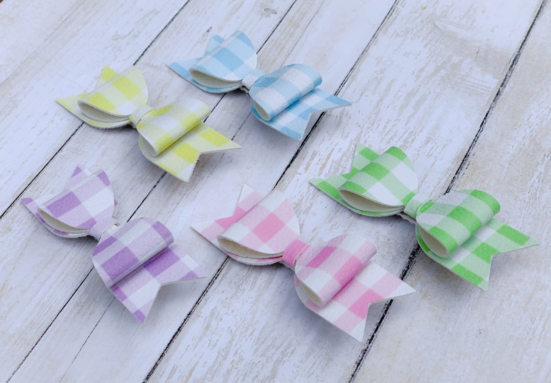 May include: Five gingham hair bows in different pastel colours. The bows are made of fabric with a white and coloured checkered pattern. The colours are light blue, yellow, green, pink, and purple.