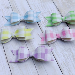 May include: Six gingham hair bows in various pastel colours, including light blue, yellow, green, pink, and purple. The bows are arranged on a white wooden surface.