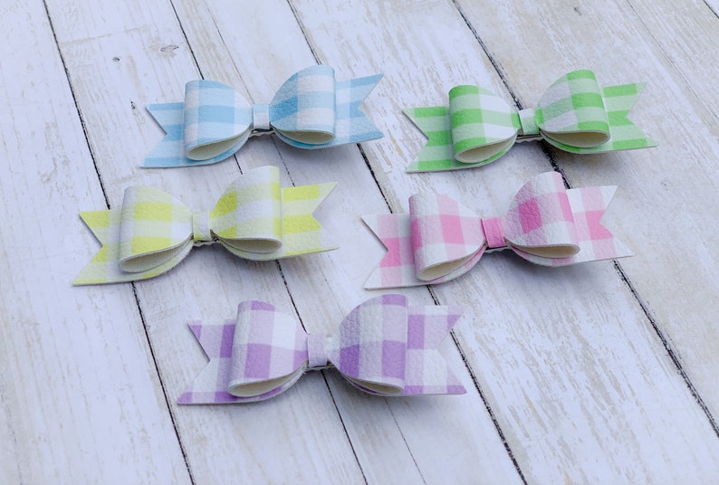May include: Six gingham hair bows in various pastel colours, including light blue, yellow, green, pink, and purple. The bows are arranged on a white wooden surface.