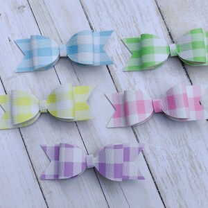 May include: Six gingham patterned hair bows in various pastel colours, including light blue, yellow, green, pink, and purple. The bows are arranged in two rows on a white wooden surface.