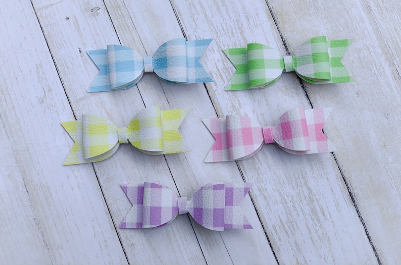 May include: Six gingham patterned hair bows in various pastel colours, including light blue, yellow, green, pink, and purple. The bows are arranged in two rows on a white wooden surface.