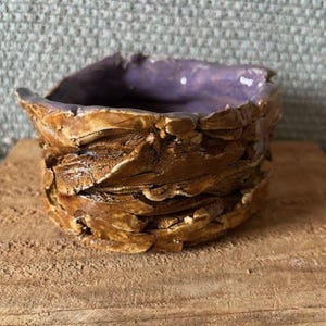 May include: A handmade ceramic bowl with a textured, layered exterior in shades of brown and tan. The interior is a glossy purple. The bowl sits on a wooden surface.