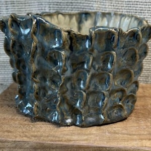 May include: A ceramic bowl with a textured, ruffled exterior featuring a variegated glaze in shades of deep blue, slate gray, and earthy brown. The rim is uneven and wavy, and the interior surface appears smooth with a lighter, cream-colored finish.