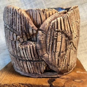 May include: A cylindrical ceramic planter with a textured, light brown exterior featuring deep vertical grooves and a cracked, weathered appearance. The vessel has a wide, open top and a dark interior, showcasing a hand-formed, layered clay construction.