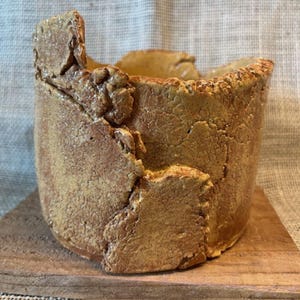 May include: A handmade, cylindrical ceramic pot with a rough, textured surface. The pot is a warm, earthy tone with a cracked, irregular rim and sides. It sits on a small, square wooden base, showcasing a rustic, artisanal aesthetic.