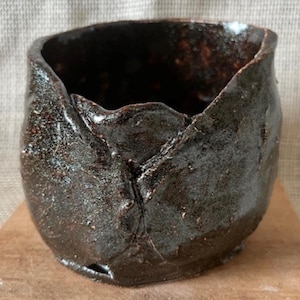 May include: A dark brown ceramic pot with an uneven, textured surface. The pot has a wide opening and a rounded shape.