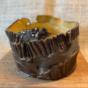 May include: A handcrafted ceramic bowl with a unique, textured design. The exterior is a dark, glossy brown, while the interior has a golden-yellow hue. The rim has an uneven, jagged edge, adding to its rustic charm. The bowl sits on a wooden surface.