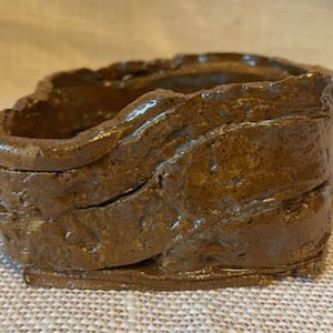 May include: A handcrafted, square-shaped brown ceramic container with a rough, textured surface. The container has a wavy rim and a hollow interior, suitable for holding small items. The earthy tones and unique design suggest a rustic aesthetic.